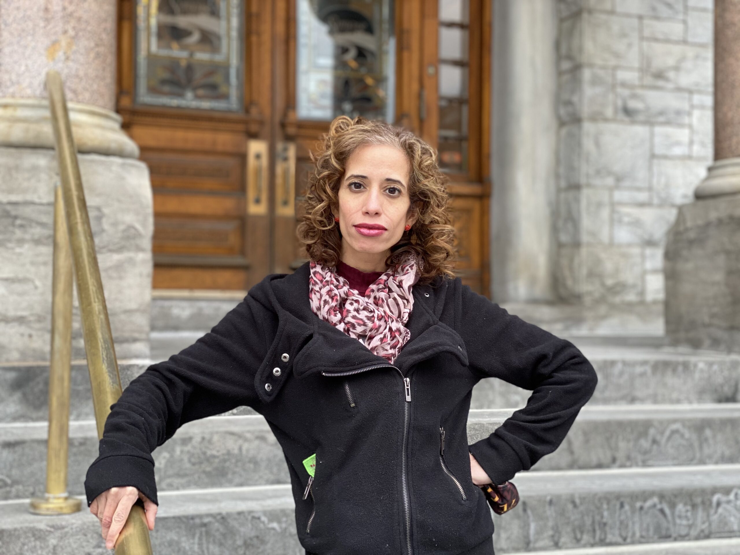Woman with a pink scark, a black coat and curly hair standing in front of city hall which has grey stone steps and brown wooden doors. 